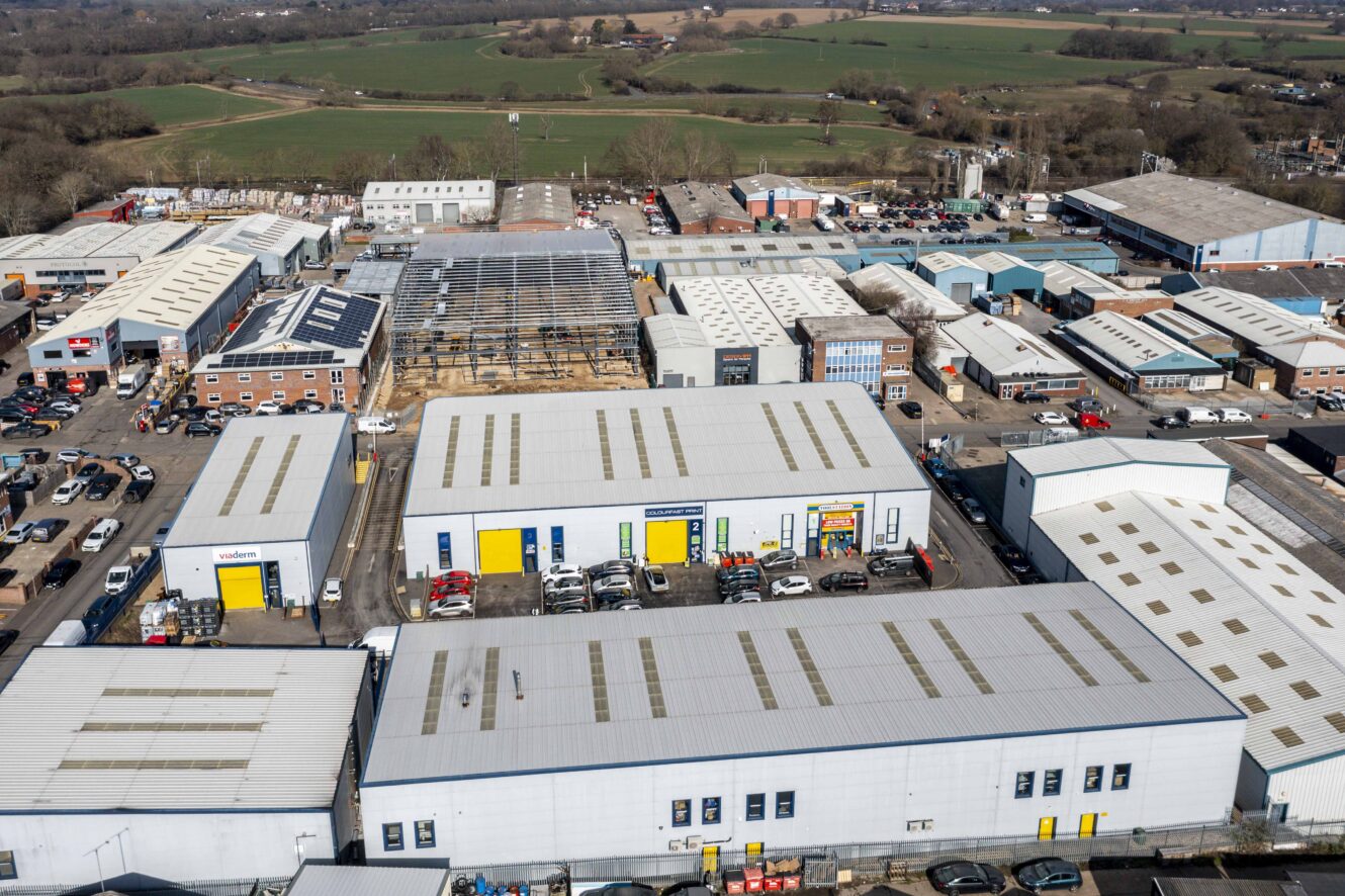Aerial view of an industrial estate with multiple warehouse buildings, parked cars, and surrounding green fields.