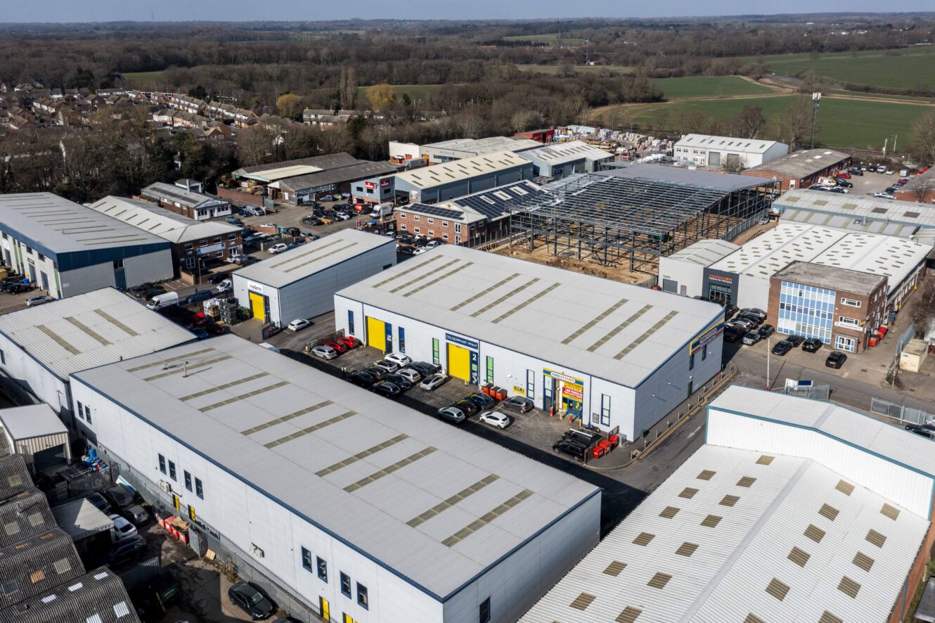 Aerial view of an industrial estate with large warehouse buildings, parking lots, and ongoing construction, surrounded by fields and residential areas.