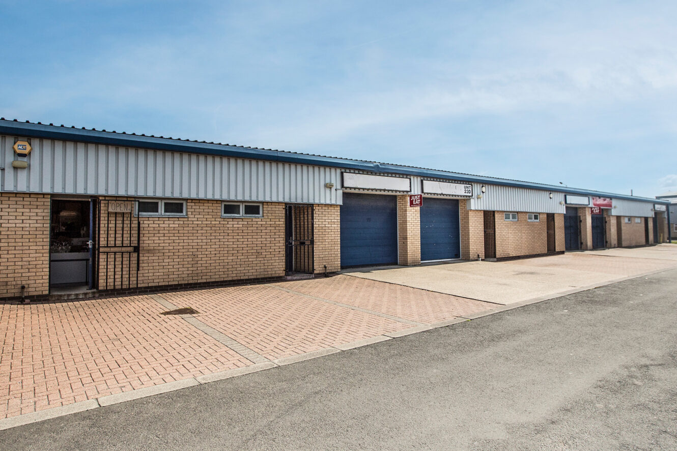 A row of light industrial units with brick walls, blue roller shutter doors, and a paved forecourt under a clear sky.