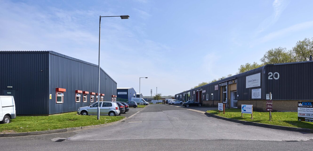 Industrial estate with commercial warehouse units on both sides, cars parked along the road, and clear blue sky overhead.