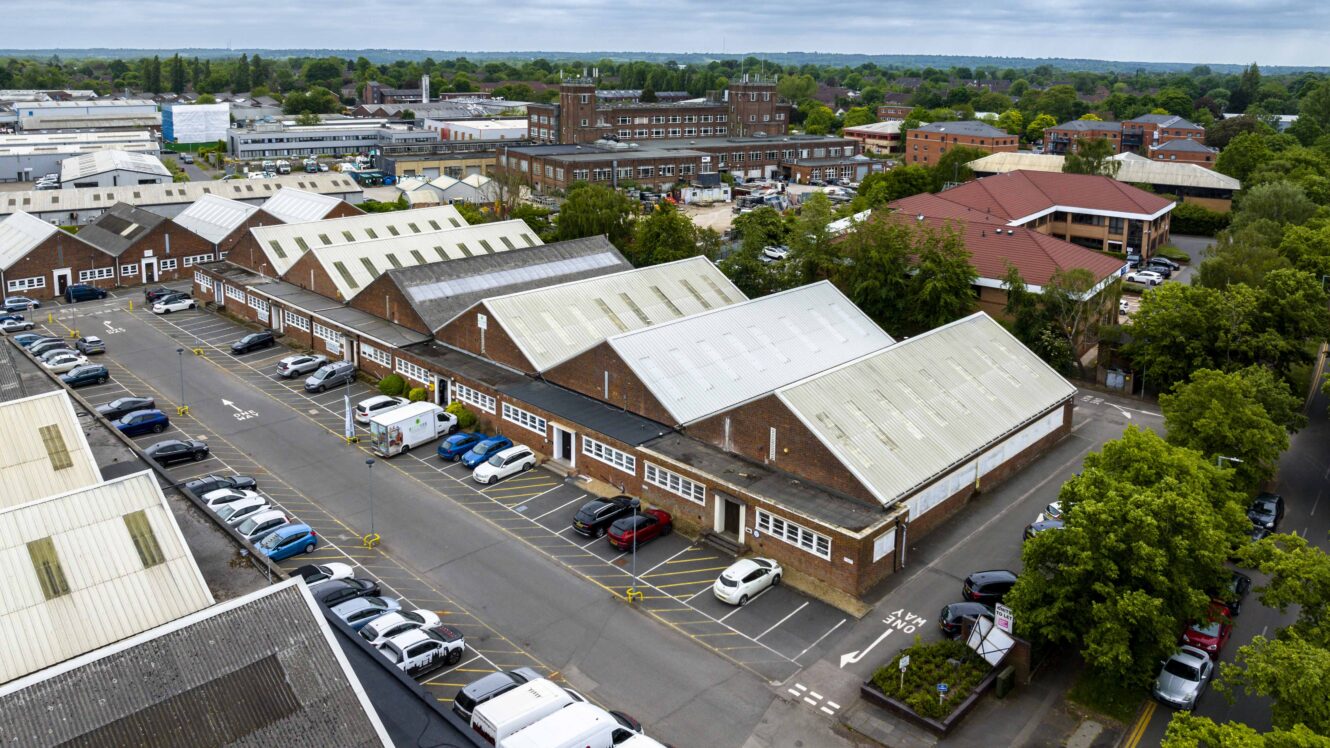 Aerial view of an industrial estate with brick warehouses, parked cars, and surrounding buildings under a cloudy sky.
