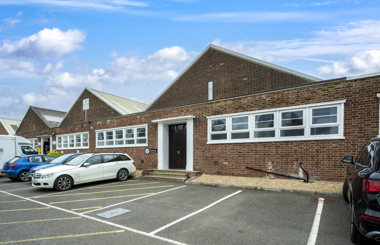 A single-story brick industrial building with sawtooth roofline, multiple windows, parked cars, and a partly cloudy sky overhead.