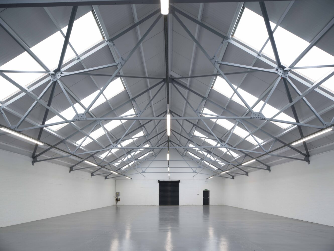 Large, empty warehouse with a high, metal truss ceiling, skylights, white walls, concrete floor, and a black roller door at the far end.