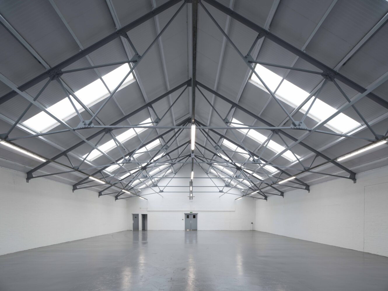 Large empty warehouse with a high pitched ceiling, exposed steel beams, and skylights. The walls are white, and there are double doors at the far end.