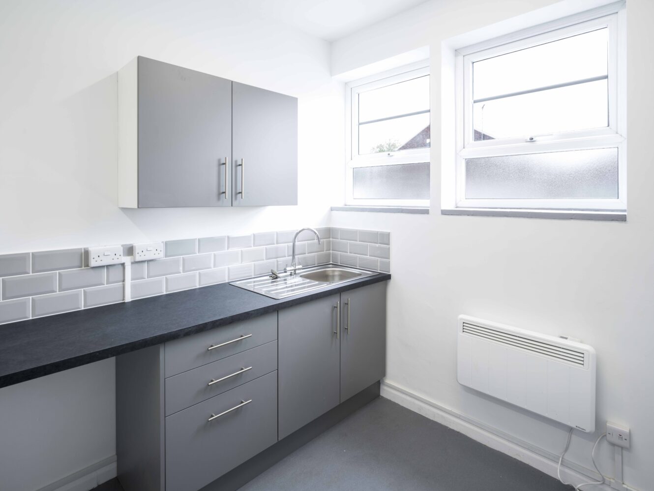 Modern kitchen with grey cabinets, a black countertop, a stainless steel sink, tiled backsplash, two windows, and a white wall-mounted heater.
