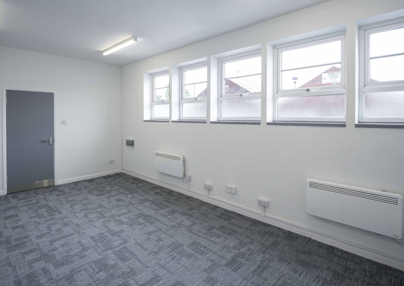 Empty room with gray carpet, white walls, four rectangular windows, two wall-mounted heaters, and a closed gray door.