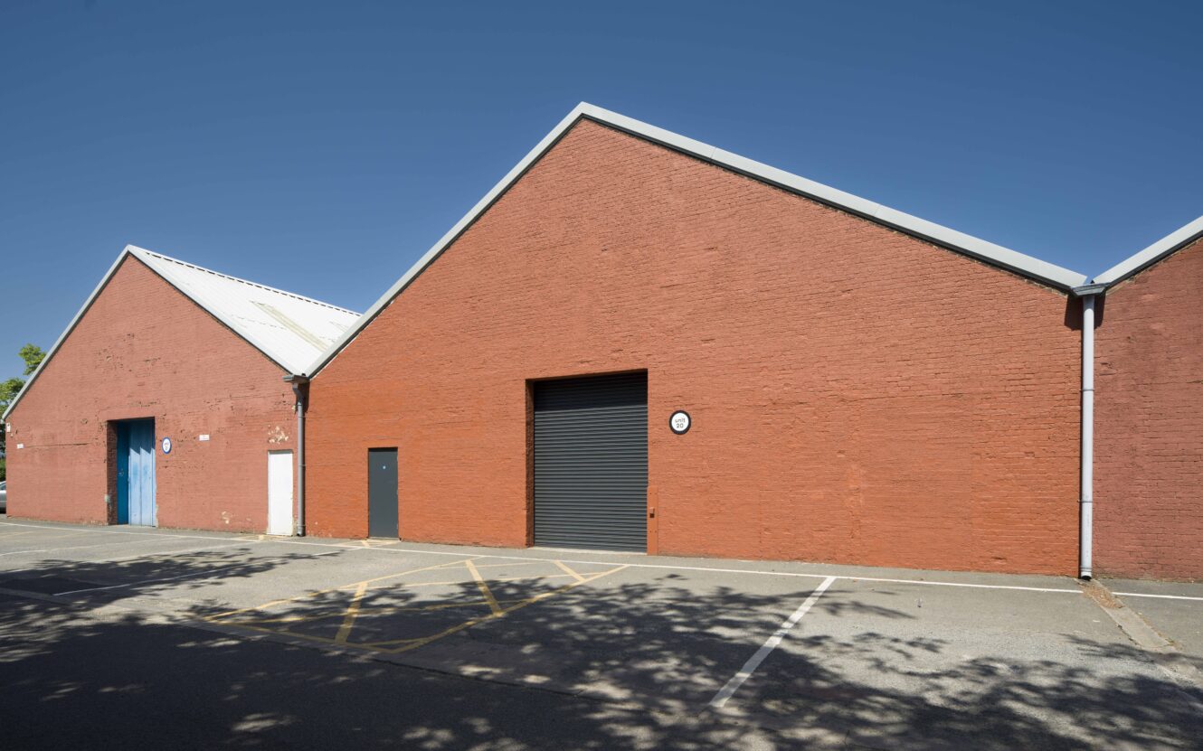 Red brick warehouse buildings with sloped roofs, featuring three doors—one blue, one gray, and one black roller shutter—next to a mostly empty parking lot under clear blue sky.