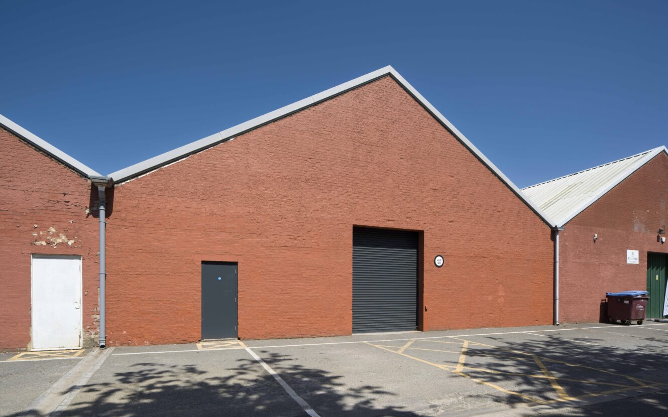 A red brick warehouse with a peaked roof, featuring a black roller door, a gray door, and a white door, with empty parking spaces in front under a clear blue sky.