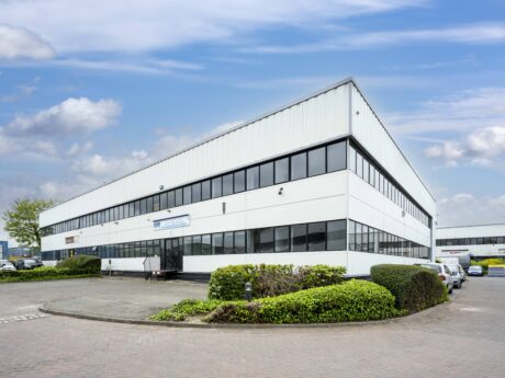 Large modern industrial or office building with white facade, black trim, large windows, and parked cars; landscaping with bushes in front; blue sky with clouds overhead.