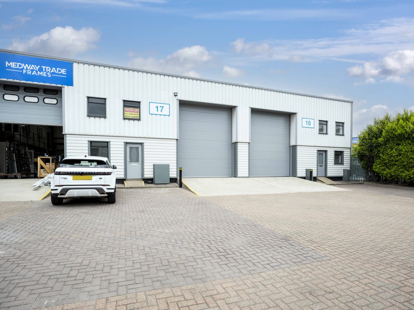 A white car is parked outside an industrial building with two large gray roller doors labeled units 17 and 18 under a blue Medway Trade Frames sign.