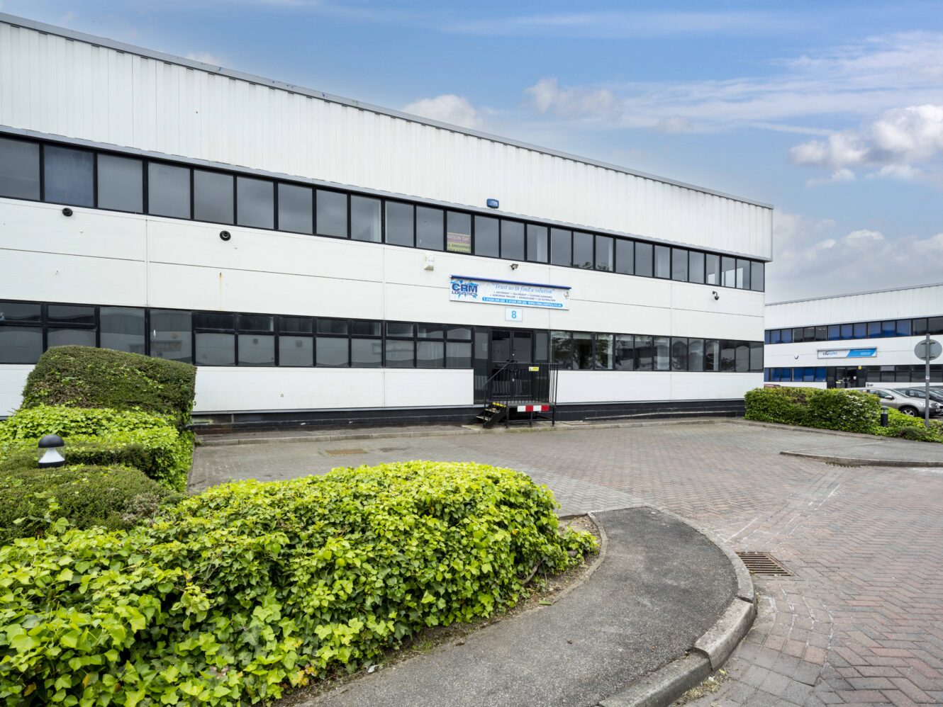 A two-story industrial office building with white paneling, black trim, and large windows, surrounded by shrubs and a paved driveway under a partly cloudy sky.