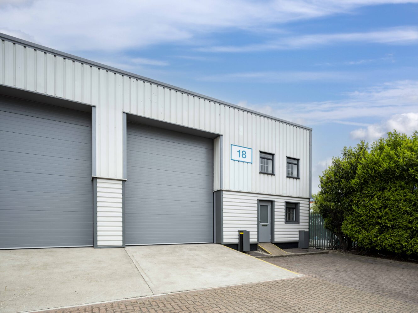 A modern industrial warehouse unit with large gray roller doors, a small entry door, and a sign displaying the number 18, set against a partly cloudy sky.