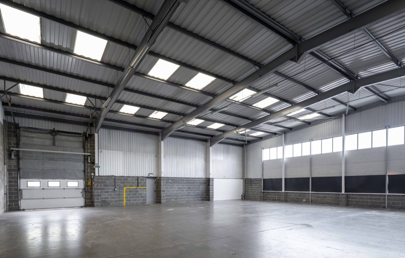 Empty industrial warehouse interior with high ceilings, large windows, concrete floor, overhead lights, and a roller shutter door.