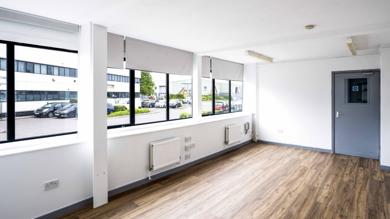 Empty office room with wood flooring, large windows with blinds, white walls, radiators, electrical outlets, and a gray door; parking lot and buildings visible outside.