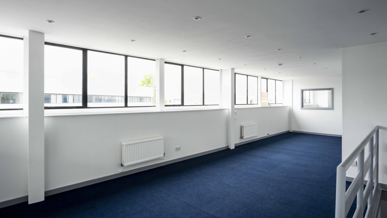 A bright, empty room with large windows, white walls, blue carpet, radiators, and a railing on the right side.