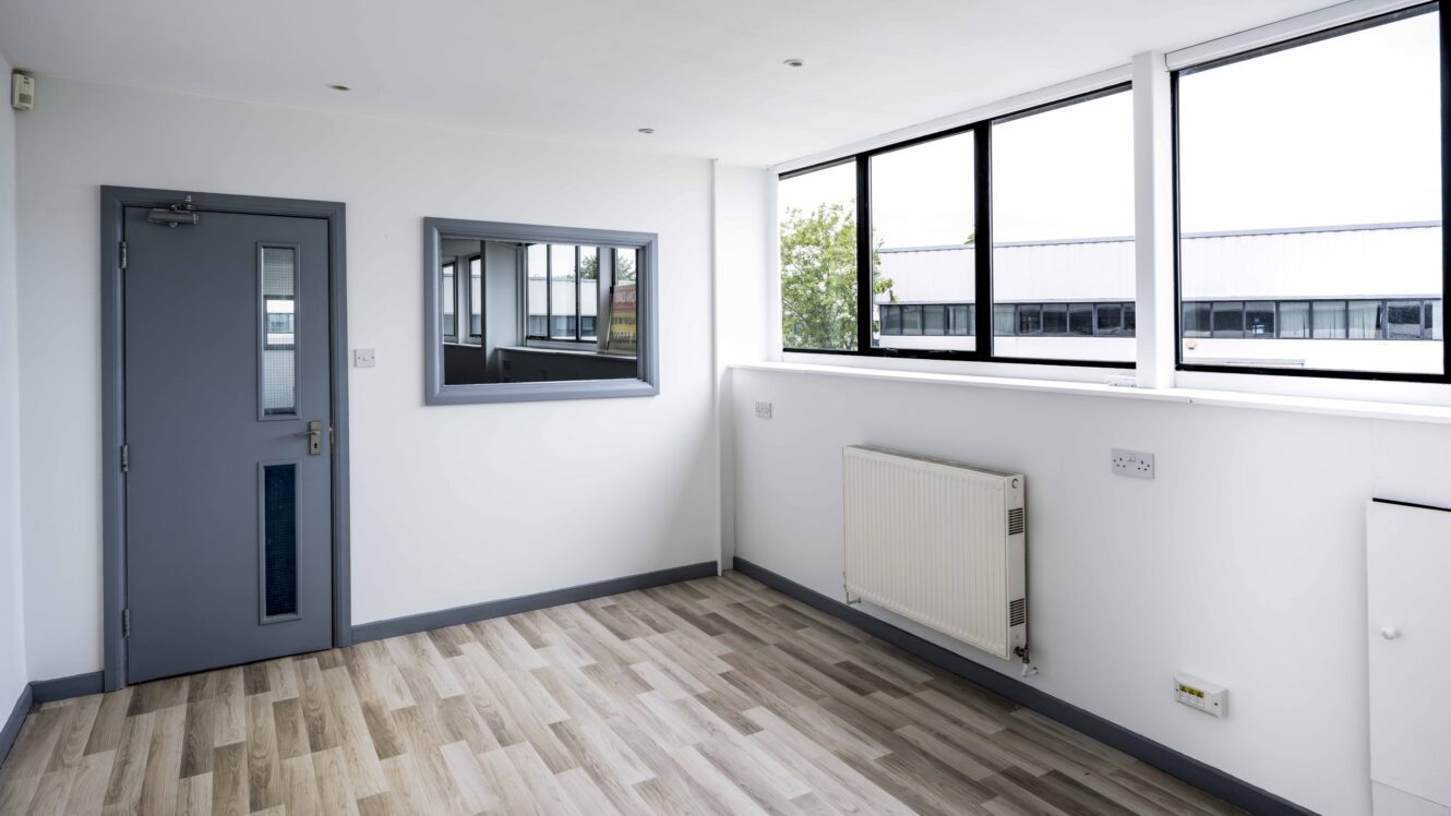 An empty, modern room with light wood flooring, white walls, large windows, a radiator, a wall mirror, and a closed gray door.