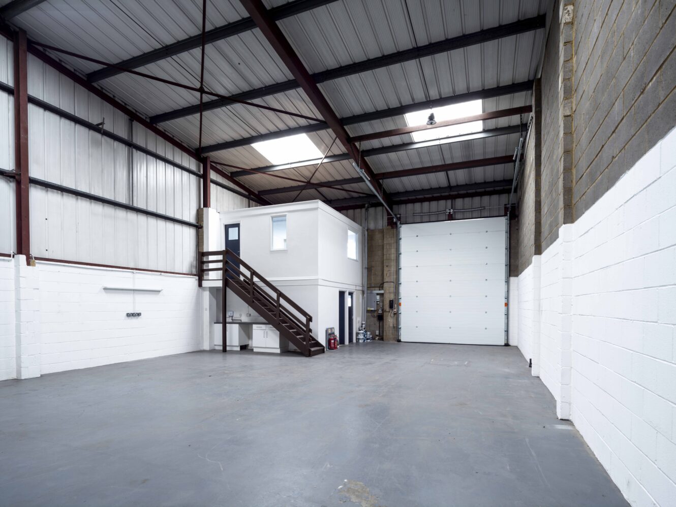 Interior of an empty industrial warehouse with high ceilings, white walls, a small office structure, staircase, and a large roller shutter door.