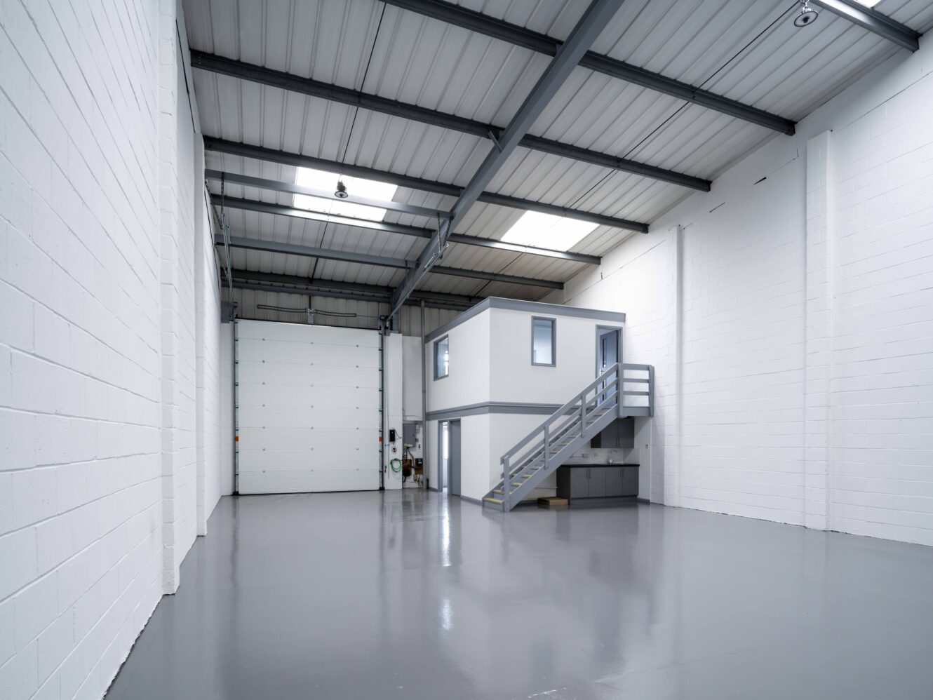 Interior of an empty warehouse unit with white walls, grey polished floor, high ceiling, and a small office structure with stairs in one corner.