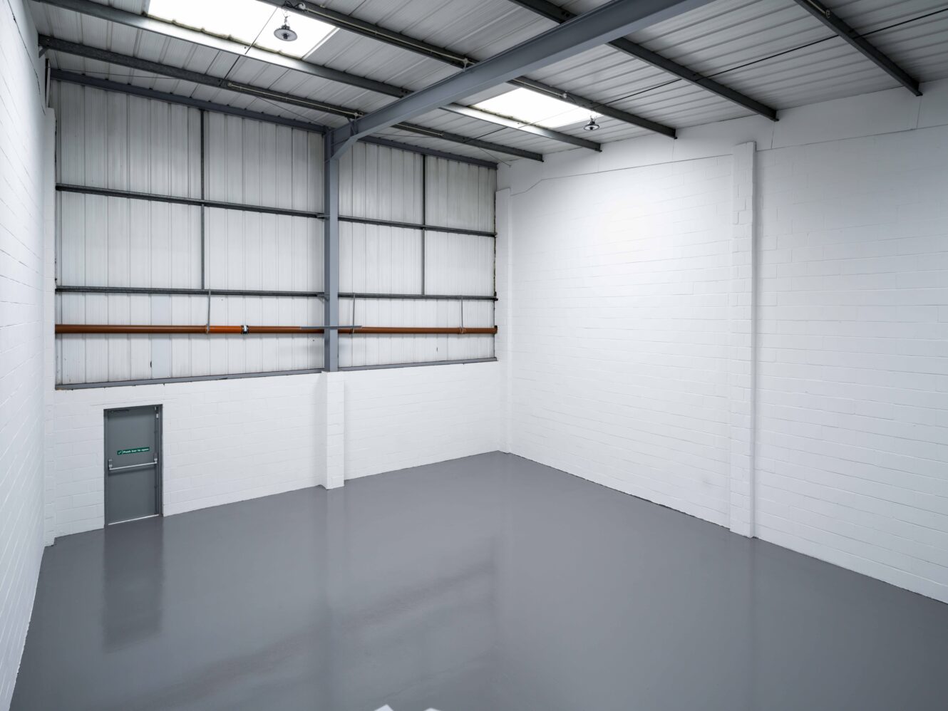 Empty industrial warehouse space with white brick walls, a gray polished floor, metal ceiling beams, and a green emergency exit door on the left wall.