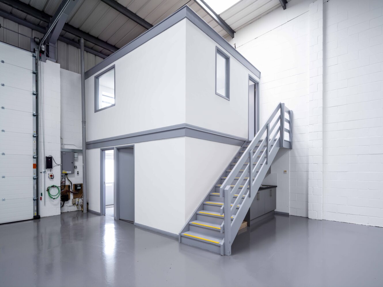 A small, modern mezzanine office with stairs inside a clean, empty industrial warehouse space.