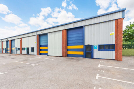 A row of industrial warehouse units with blue and yellow doors, brick accents, and numbered bays, set in a parking lot under a partly cloudy sky.