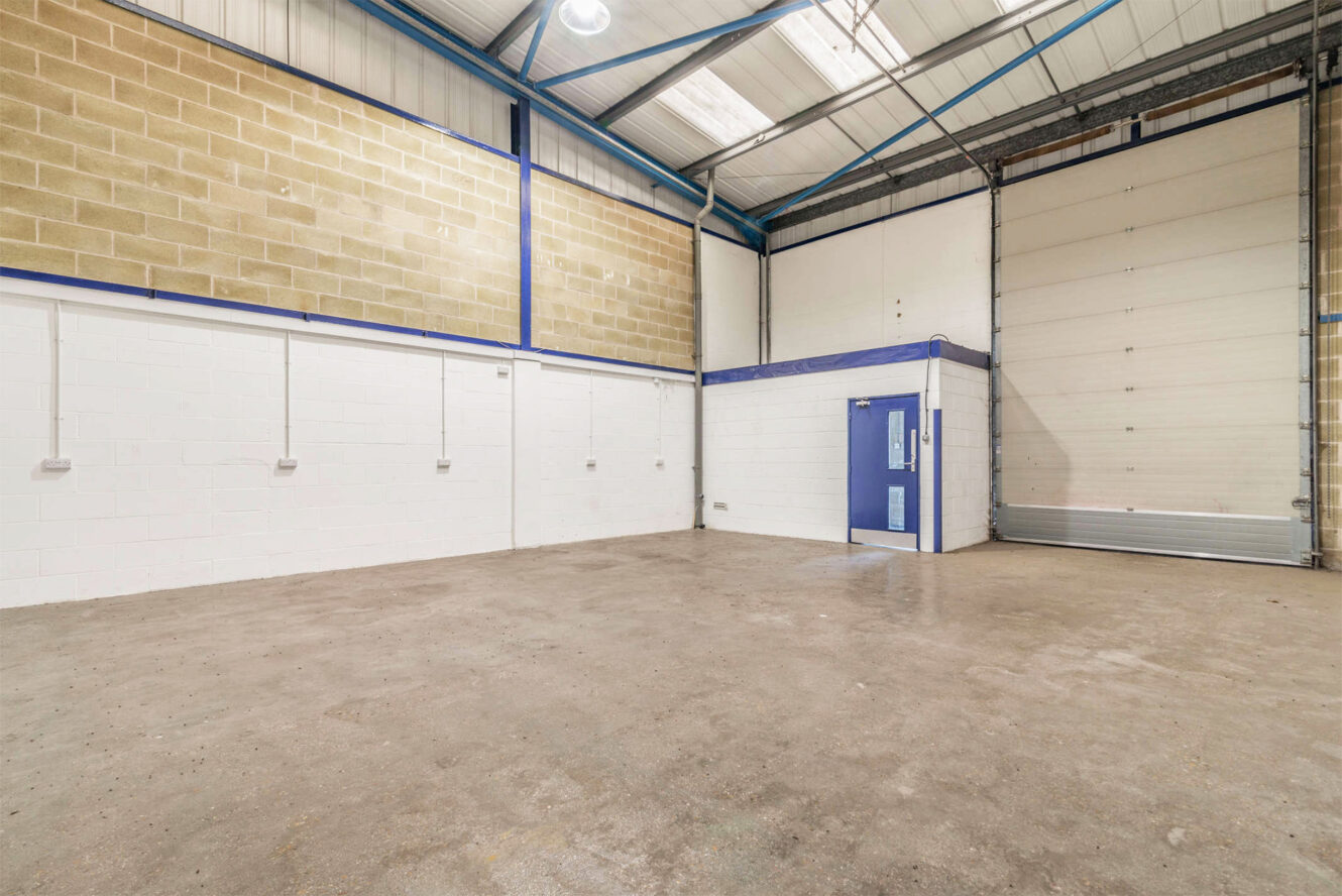 Empty industrial warehouse unit with concrete floor, white and brick walls, high ceiling, overhead lighting, blue door, and large roller shutter door.