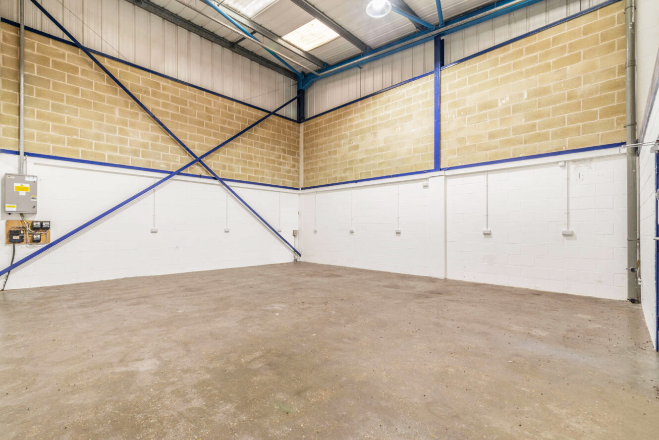 Empty industrial warehouse space with concrete floor, high ceiling, exposed brick and steel beams, white-painted lower walls, and electrical outlets along the walls.