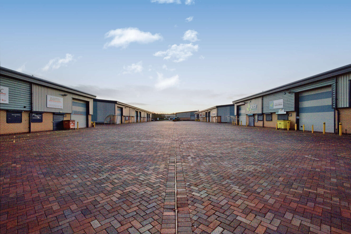 Wide view of an empty industrial park with rows of warehouse buildings on both sides and a brick-paved open area under a blue sky.