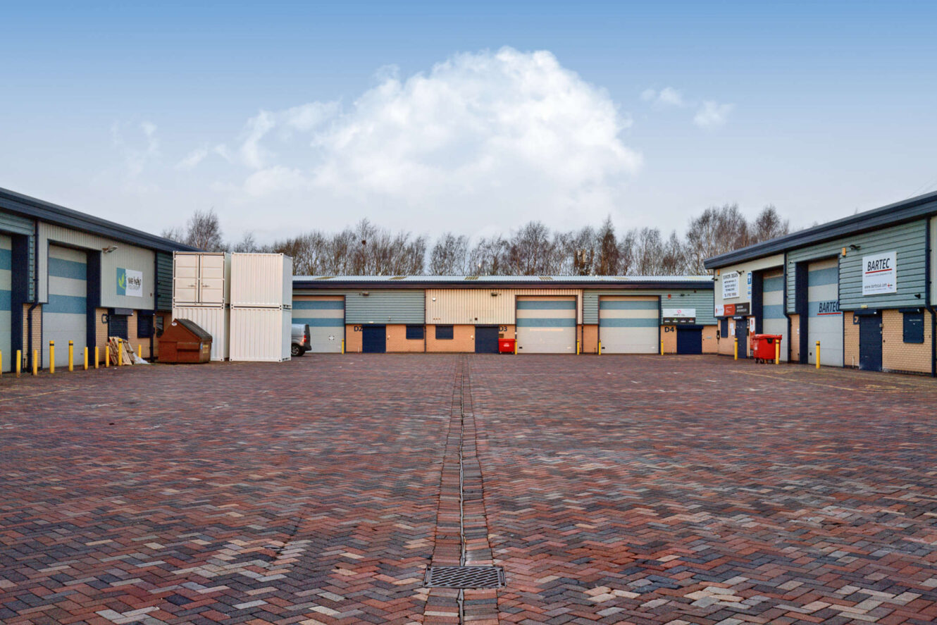 A wide, empty brick-paved lot is surrounded by industrial warehouse units with closed shutters and signs under a blue sky with scattered clouds.