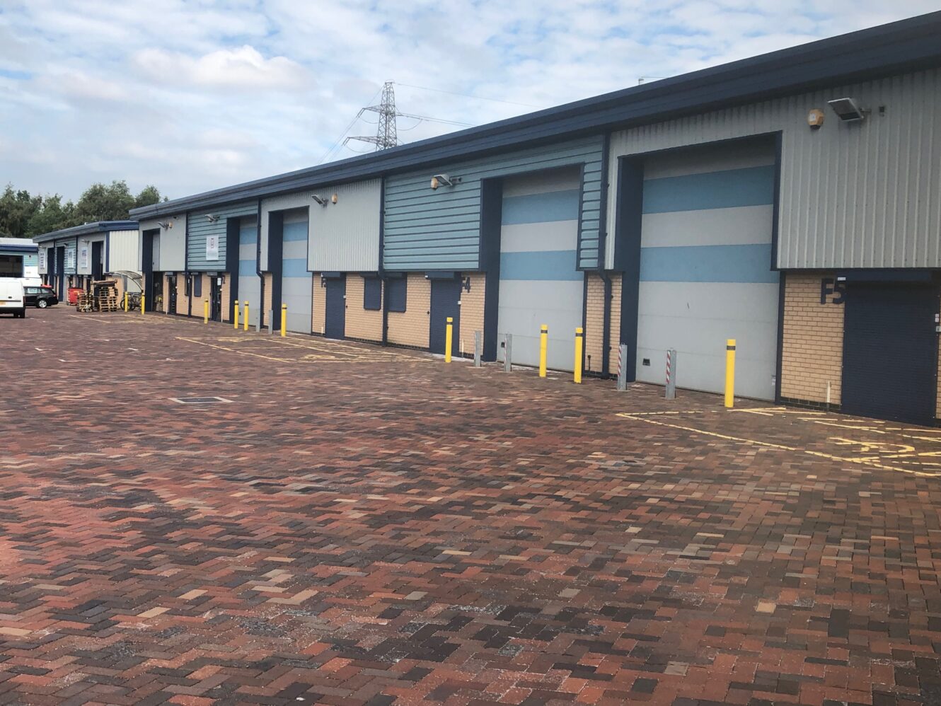 A row of industrial warehouse units with closed roller doors, brick-paved driveway, yellow bollards, and a partially cloudy sky above.