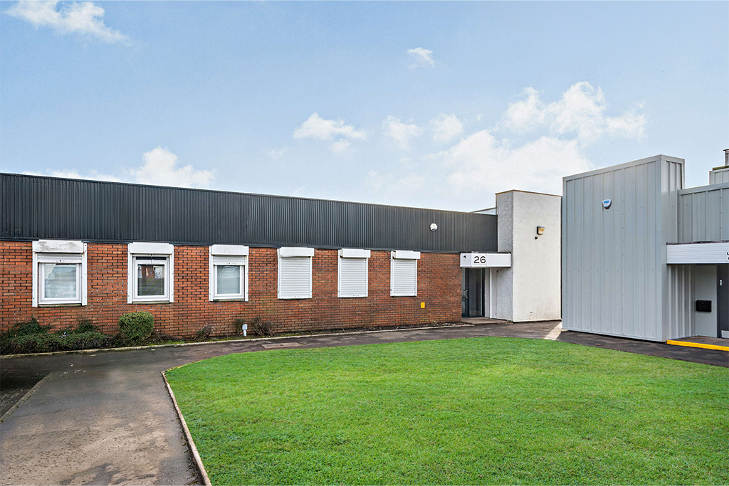 Single-story commercial or industrial building with red brick and grey metal exterior, small windows, and a neatly maintained lawn in front.