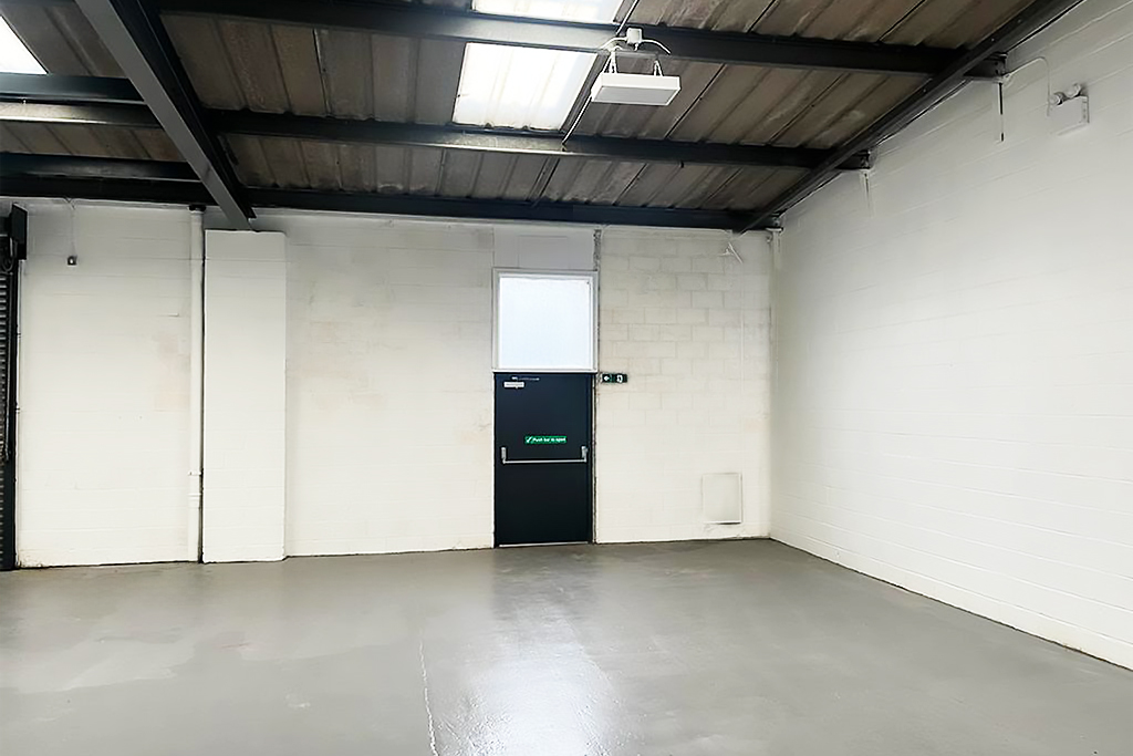 Empty industrial room with white painted brick walls, a gray concrete floor, exposed ceiling beams, fluorescent lighting, and a black emergency exit door with a green sign.