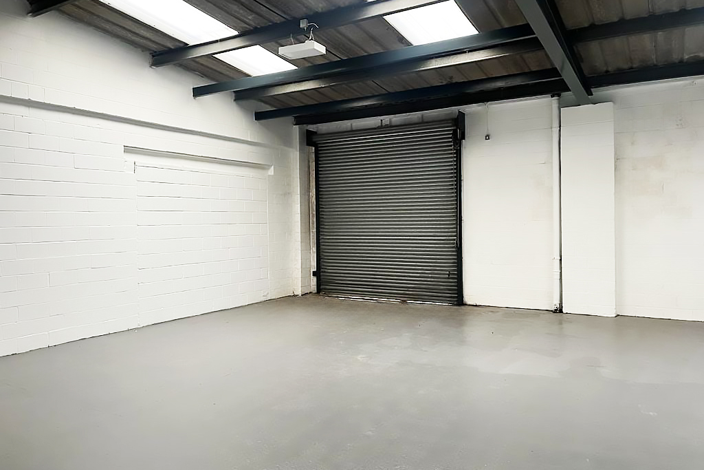 Empty industrial garage with white brick walls, concrete floor, exposed beams, skylights, and a large metal roll-up door.