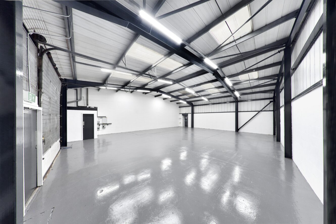 Interior of an empty industrial warehouse with polished grey floor, white walls, exposed beams, and overhead lighting.