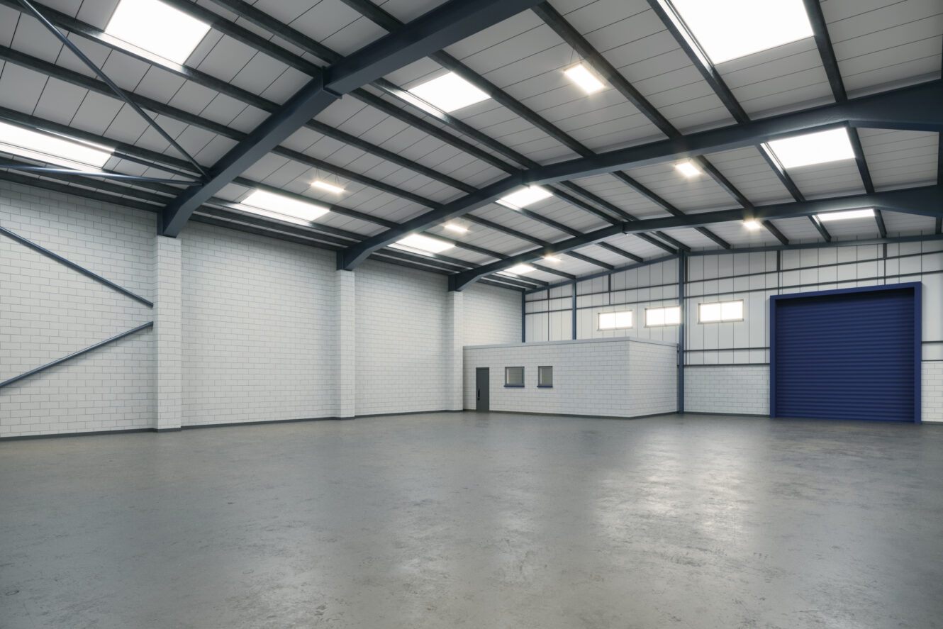 Interior of an empty industrial warehouse with high ceilings, white brick walls, skylights, a blue roller door, and a small office area inside.