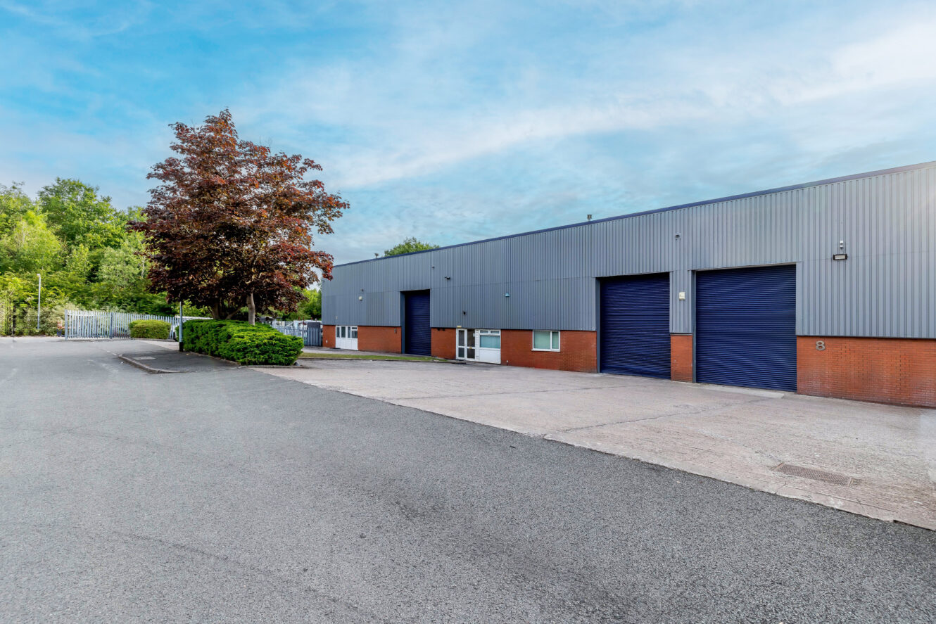 Industrial warehouse building with blue doors, brick and metal exterior, surrounded by trees and an empty asphalt parking area under a clear sky.