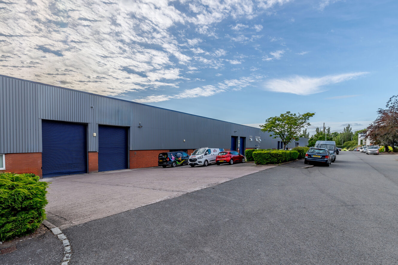 A modern industrial building with blue garage doors, cars parked in front, and a clear sky above. Bushes line the edge of the parking area.