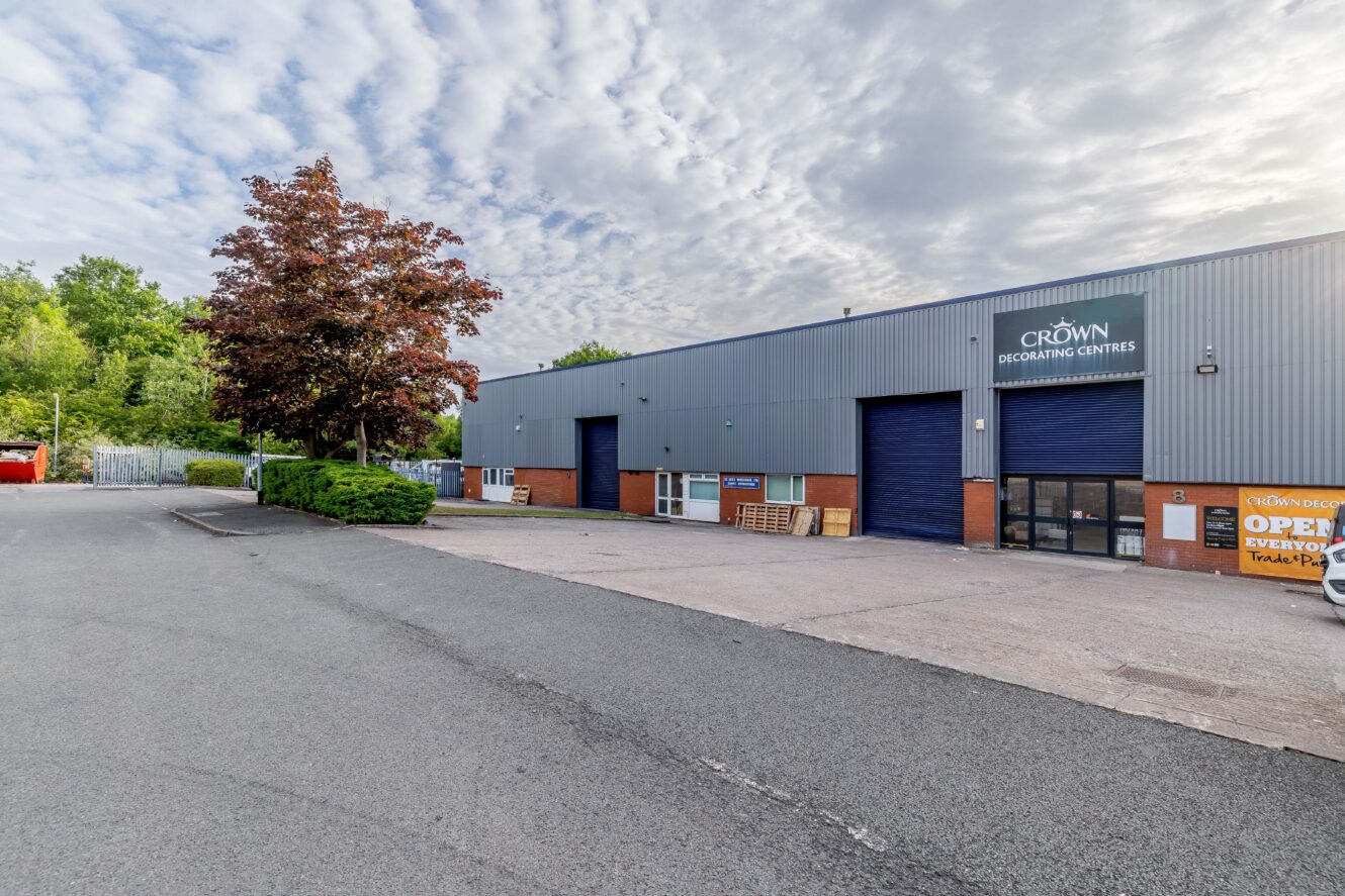 A large industrial building with blue roller doors and a Crown Decorating Centres sign, surrounded by a parking lot and some greenery under a partly cloudy sky.