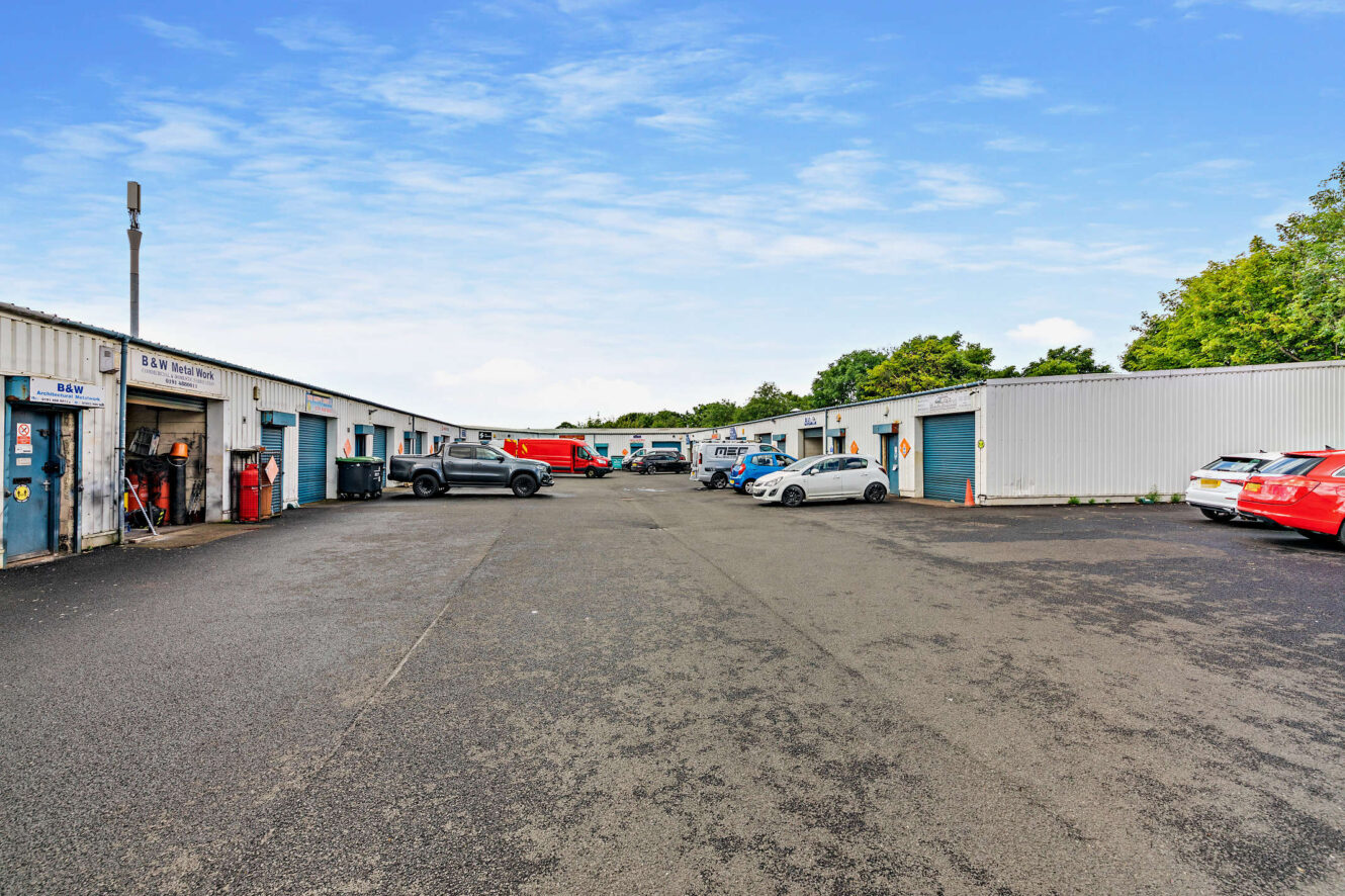 Industrial estate with several single-story warehouse units and parked vehicles on both sides of a wide asphalt lot under a partly cloudy sky.