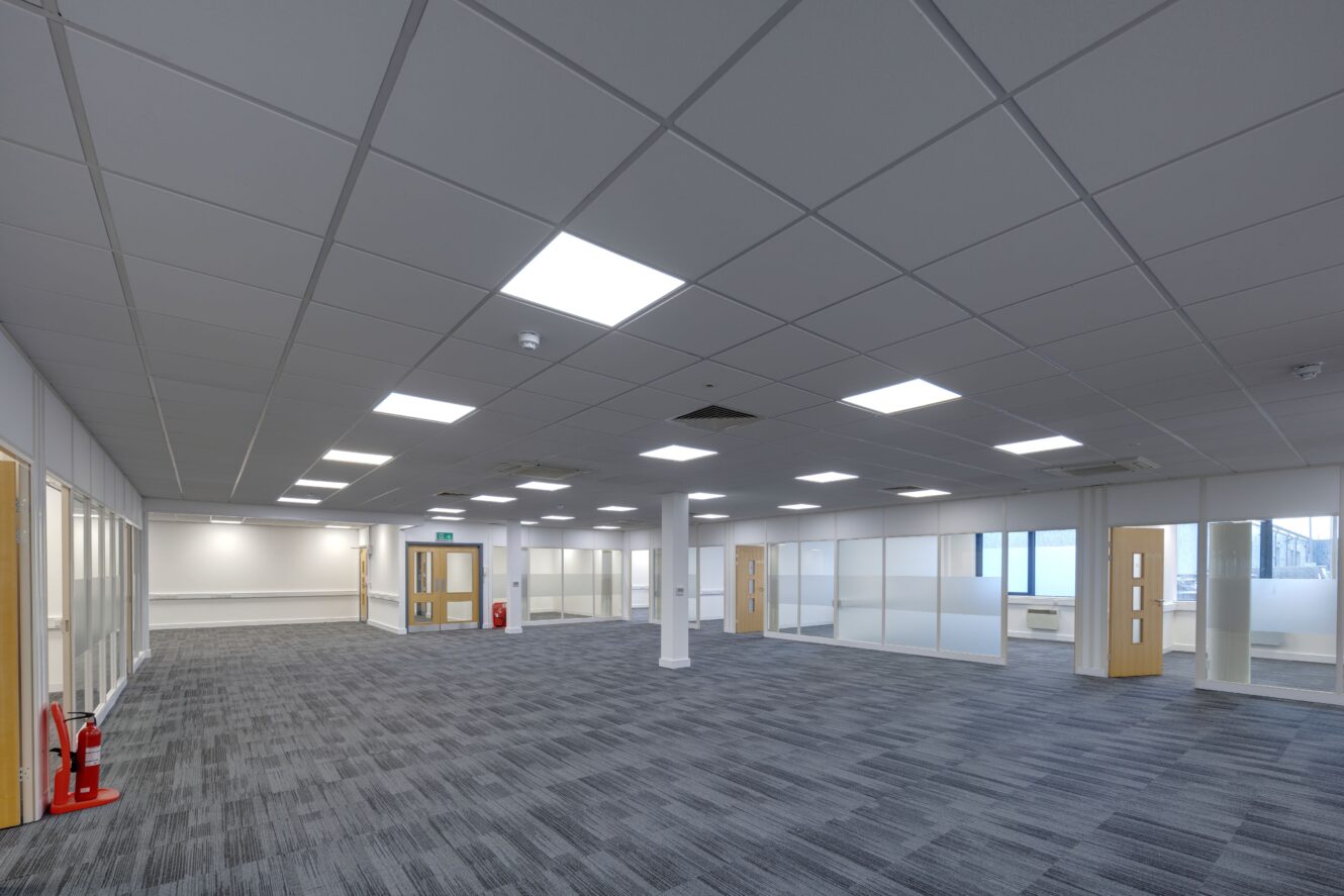 Large empty office space with grey carpet, white ceiling tiles, fluorescent lighting, glass-walled rooms, and wooden doors; two fire extinguishers are visible.