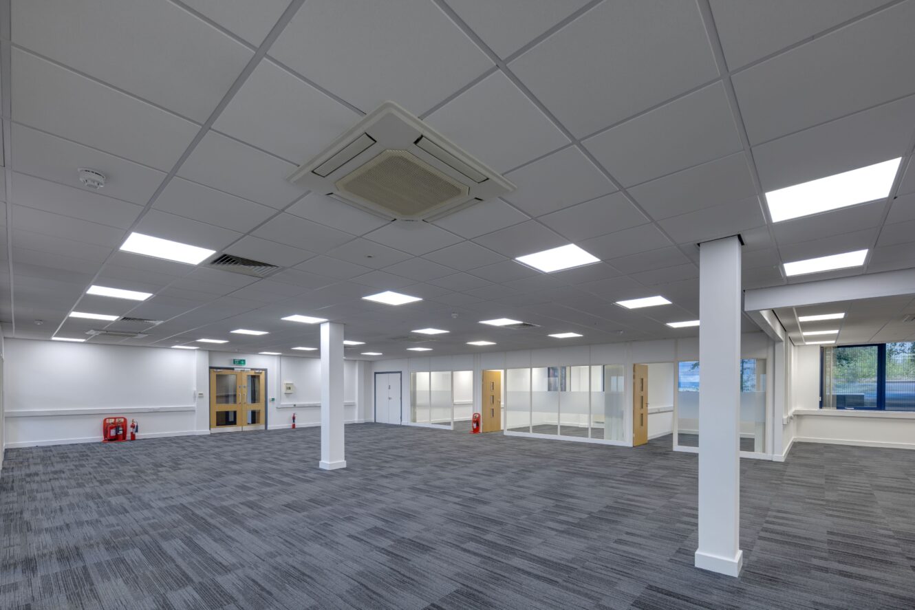 Empty modern office space with gray carpet, white walls, ceiling lights, glass partitions, and fire extinguishers near the doors.