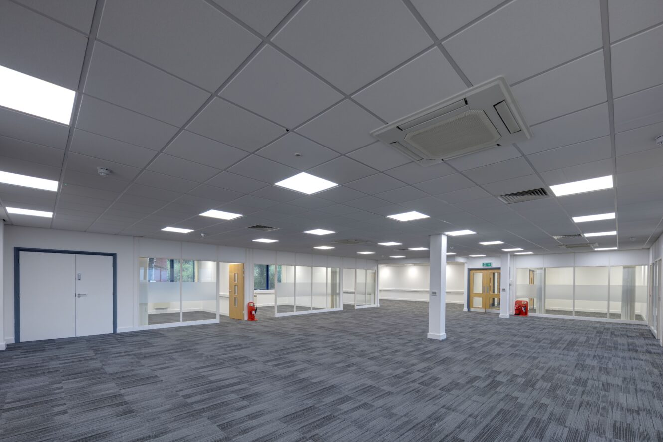 Large, empty office space with gray carpet, white walls, glass partitions, and fluorescent ceiling lights. Fire extinguishers are visible near doors and windows along the walls.