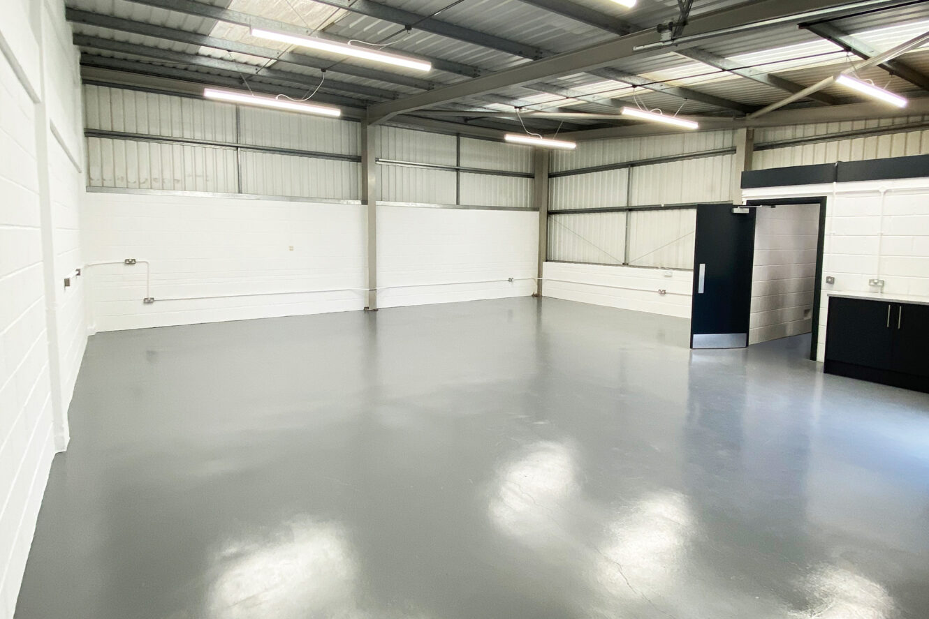 Empty industrial room with white painted brick walls, gray polished concrete floor, exposed metal ceiling, and fluorescent lighting. A black door and small sink area are visible on the right.