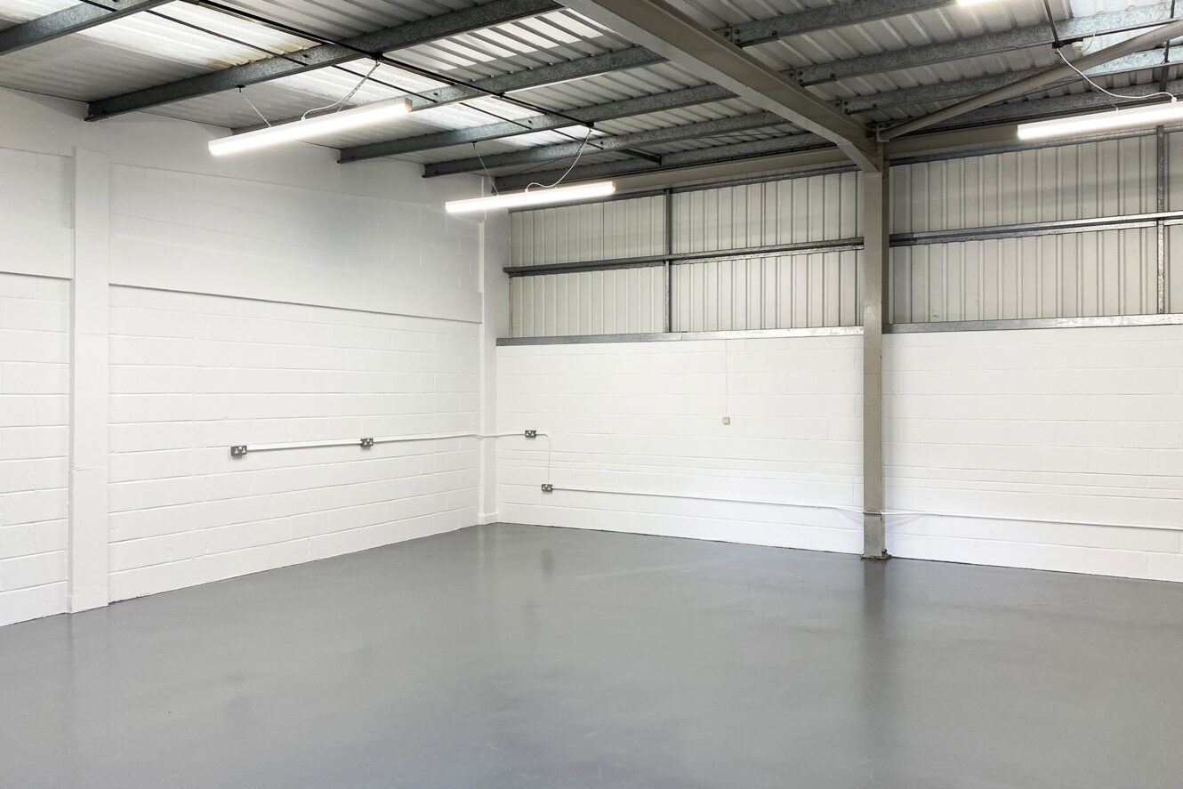 Empty industrial warehouse unit with white painted brick walls, grey concrete floor, exposed metal ceiling, and fluorescent strip lighting.