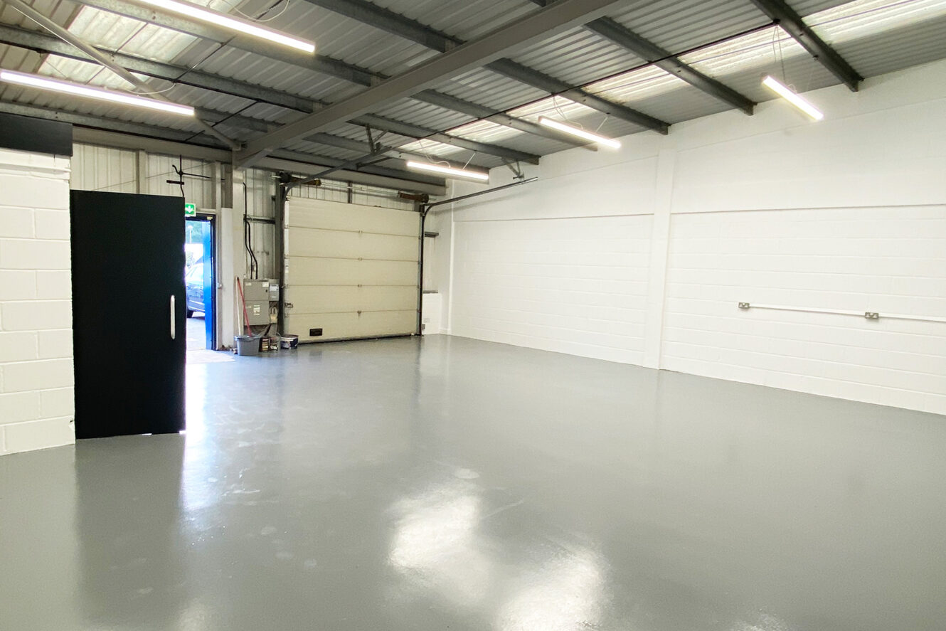 An empty industrial warehouse with gray flooring, white brick walls, exposed ceiling beams, fluorescent lights, a large garage door, and a black entry door.
