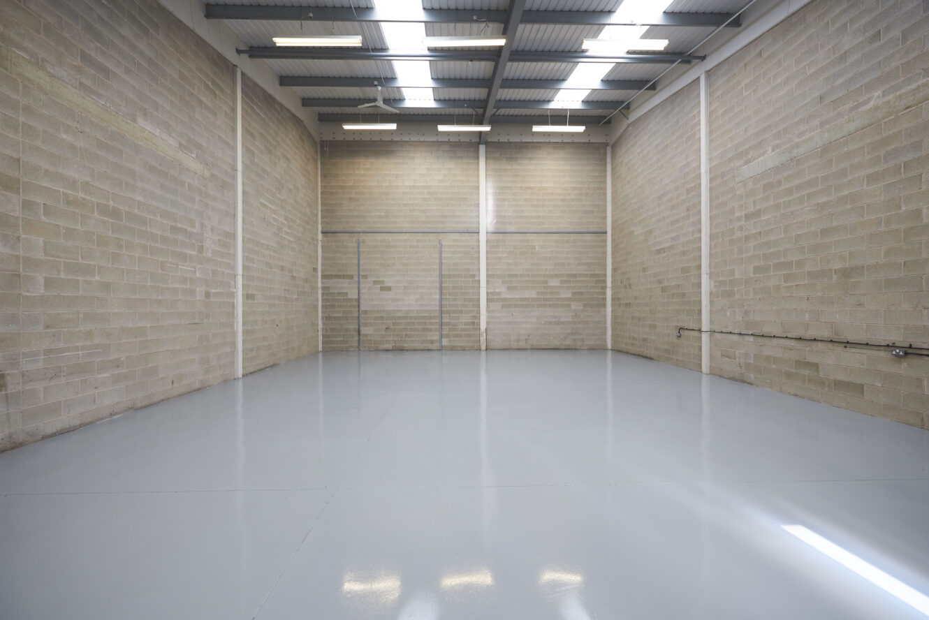 An empty industrial warehouse unit with high ceilings, exposed brick walls, polished concrete floor, and fluorescent lighting overhead.