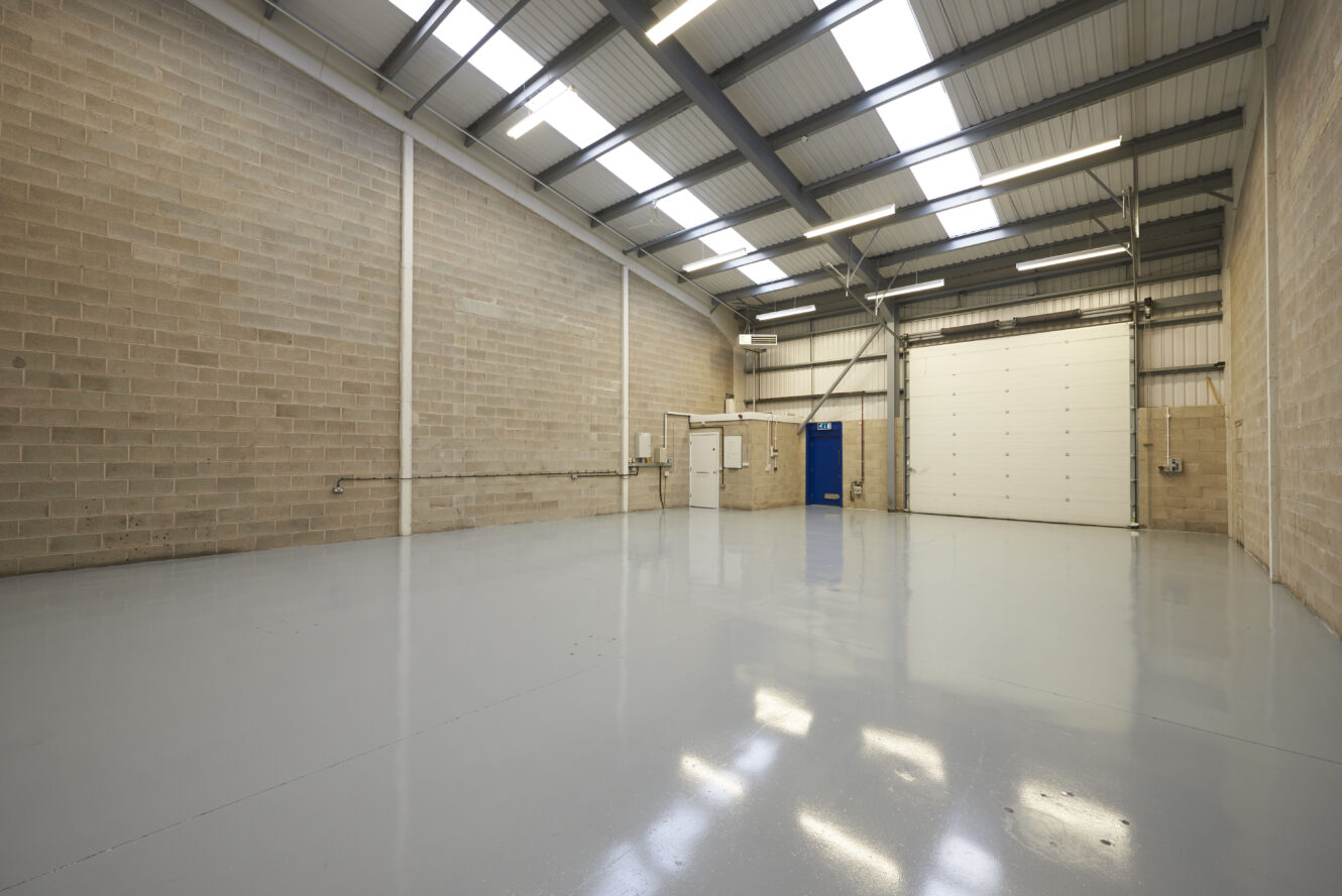 Empty industrial warehouse with polished concrete floor, high ceiling, beige brick walls, skylights, and a large white roller shutter door.