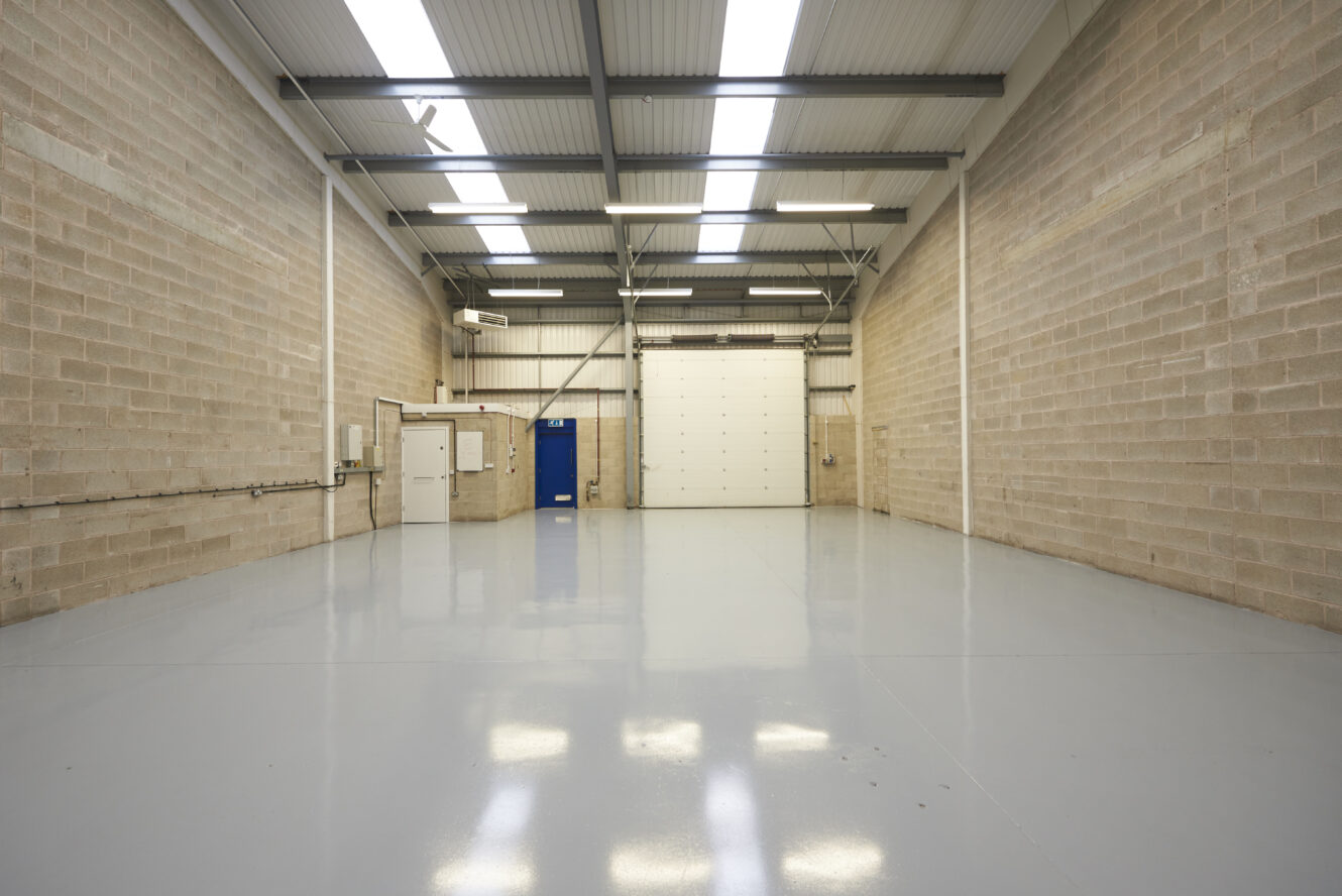 Empty industrial warehouse with bare brick walls, polished concrete floor, high ceiling, fluorescent lighting, and large white garage door at the far end.