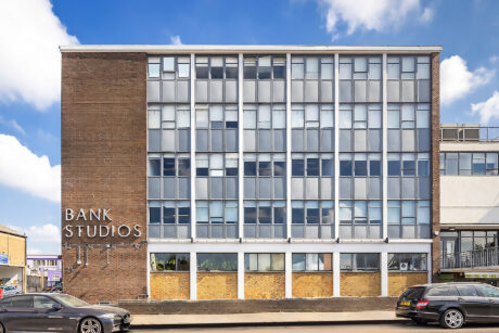 A four-story office building labeled BANK STUDIOS on its brick facade, with large grid windows and cars parked on the street in front.