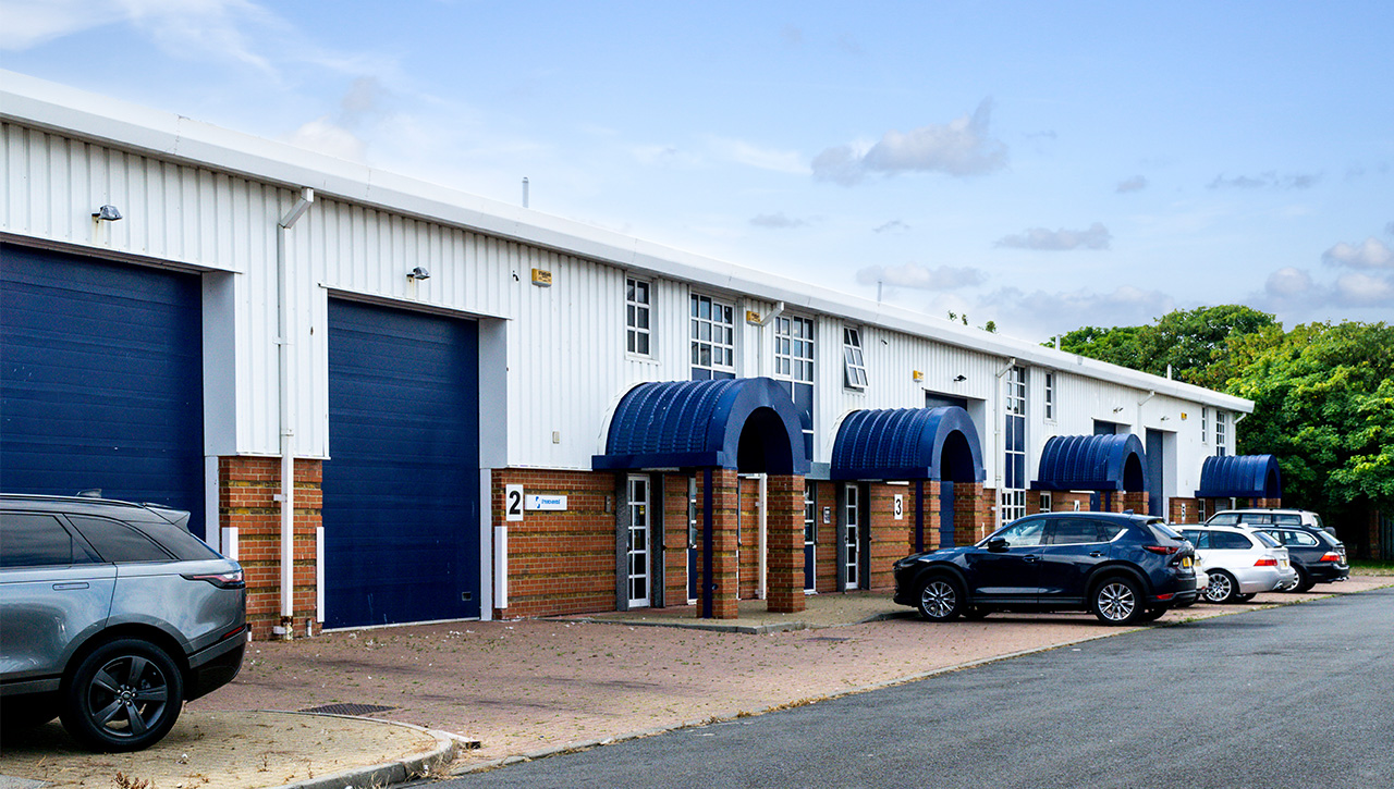 A row of industrial warehouse units with blue awnings and roller doors, several cars parked in front, and trees in the background.